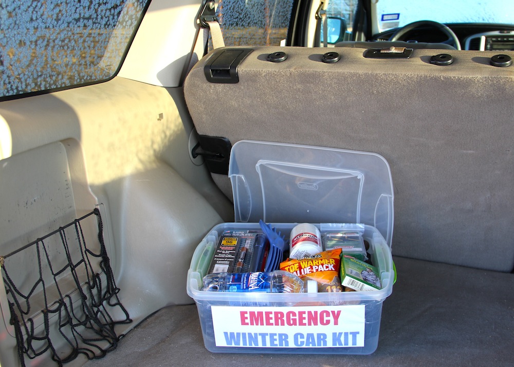 A car parked on the shoulder of a snowy road with the trunk open, showing a winter emergency kit and a folded blanket
