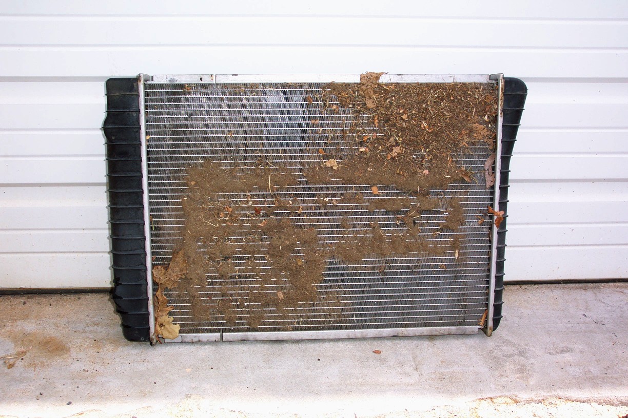 A close-up photo of a car radiator and AC condenser fins with visible trapped leaves and bugs behind the front grille