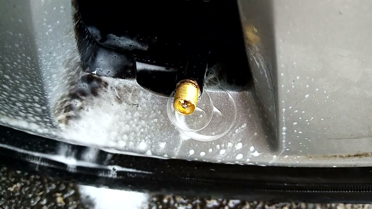 A hand sprays soapy water onto a tire valve stem to check for bubbles that indicate an air leak on a parked car