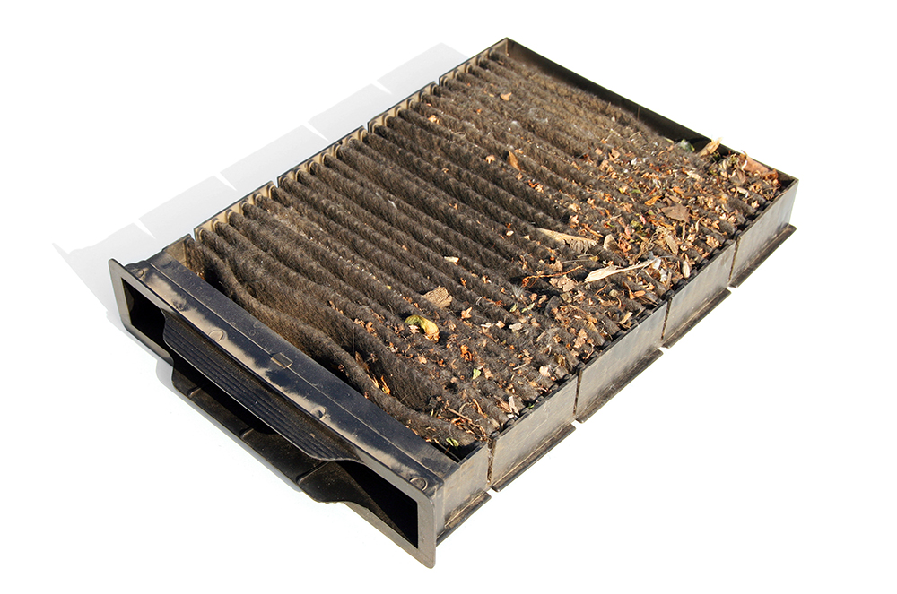 A heavily clogged cabin air filter covered in gray dust and small leaf debris being held in a person's hands