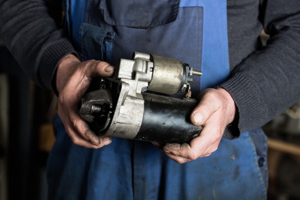 A mechanic reaching down into an engine bay toward a starter motor mounted low on the engine, with visible wiring connected to the starter