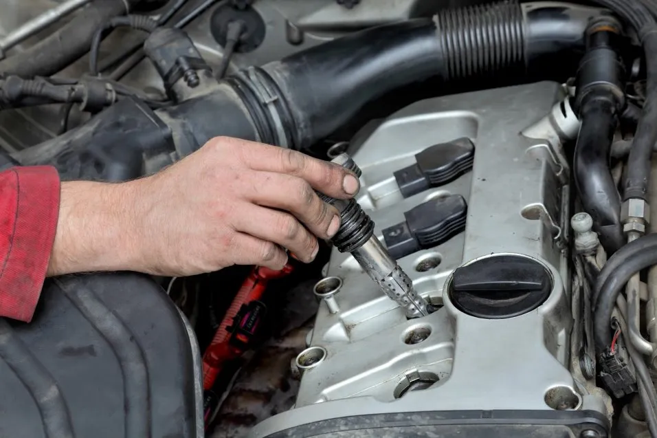 A mechanic's hand holding a modern ignition coil above an open engine bay with the spark plug well visible