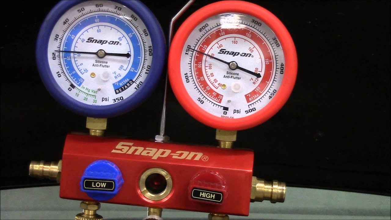 A photo of a technician connecting manifold gauge hoses to a car air conditioning service ports under the hood