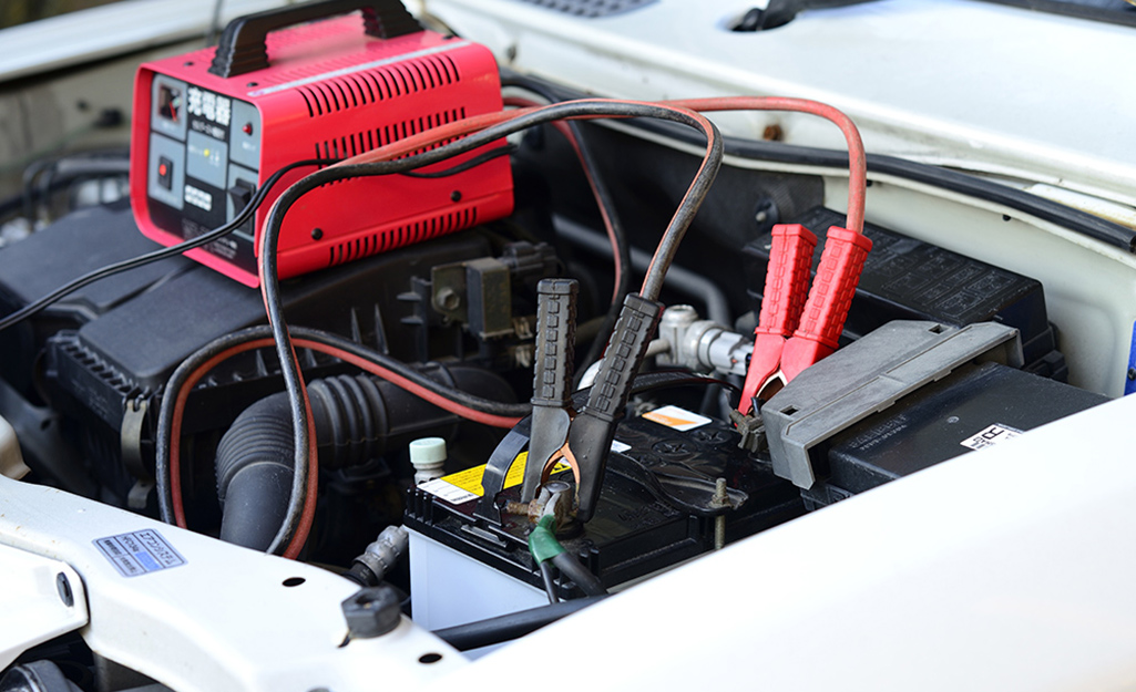 A portable battery charger connected to a car battery in an engine bay with the hood open in a residential driveway