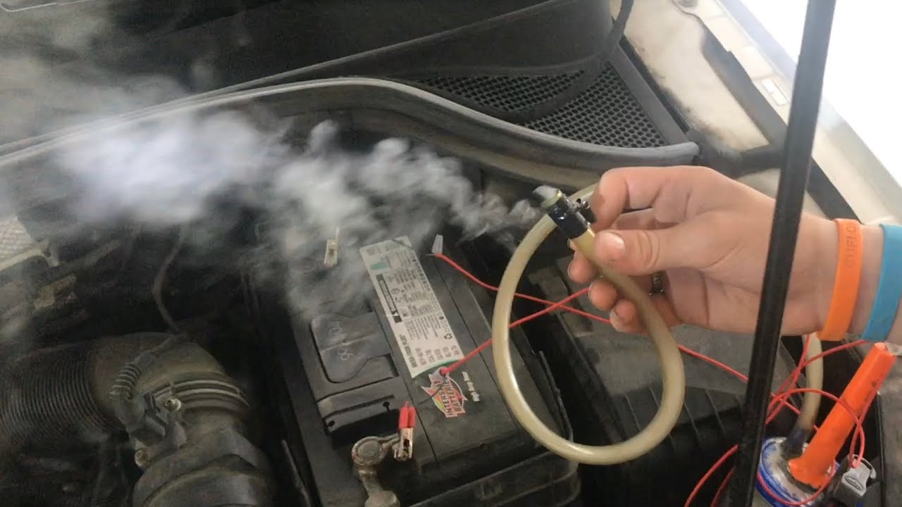 A technician performing a smoke test on a car intake system with smoke visible near the intake plumbing under the hood