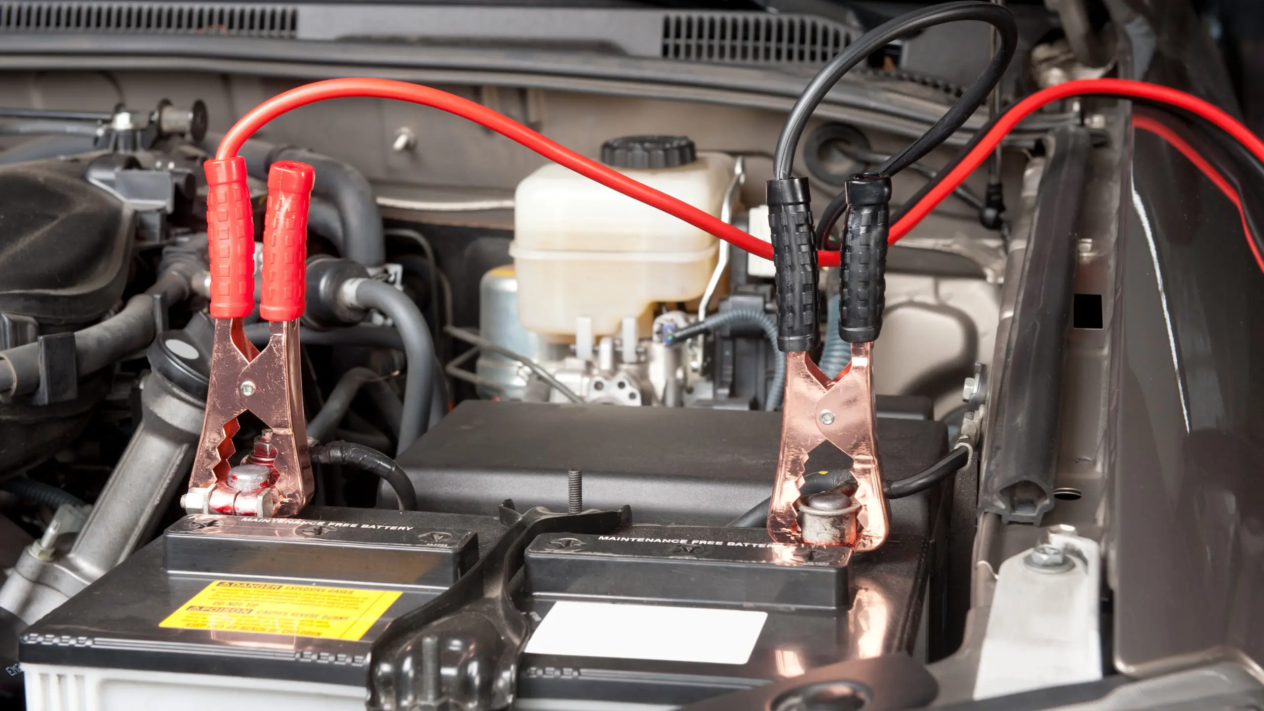 Jumper cables clipped to a car battery in an engine bay, with the red clamp on the positive terminal and the black clamp positioned at a grounding point away from the battery
