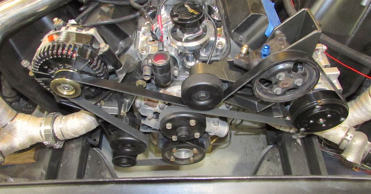 Running engine bay with a multimeter connected to the battery terminals and the alternator and serpentine belt visible in the background